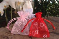 Two organza pouches with heart patterns, one pink and one red, displayed outdoors with a blurred jar in the background.